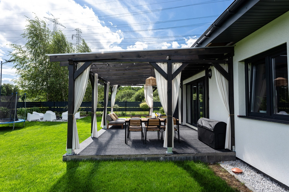 Drewniana pergola z białymi zasłonami nad tarasem z meblami ogrodowymi